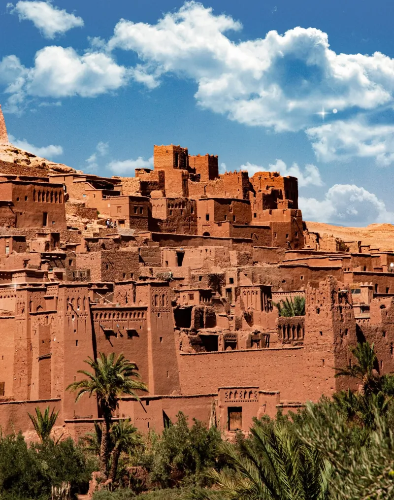 Earthen kasbah towers of Aït Benhaddou stacked up the hillside under a blue sky with clouds