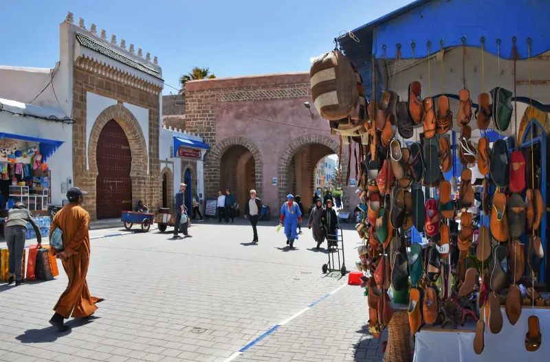 Sunny square in the Essaouira medina with a blue-and-white gateway and a stall of leather slippers