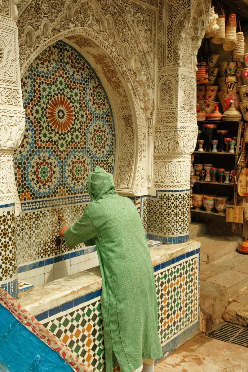 Man in a green djellaba at a zellige-tiled public fountain in the Fez medina