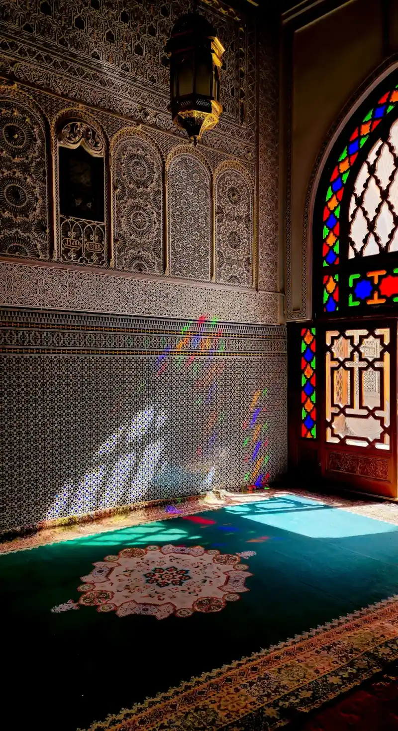 Coloured light from a stained-glass window falling across zellige tiles and carved plaster inside a Fez riad