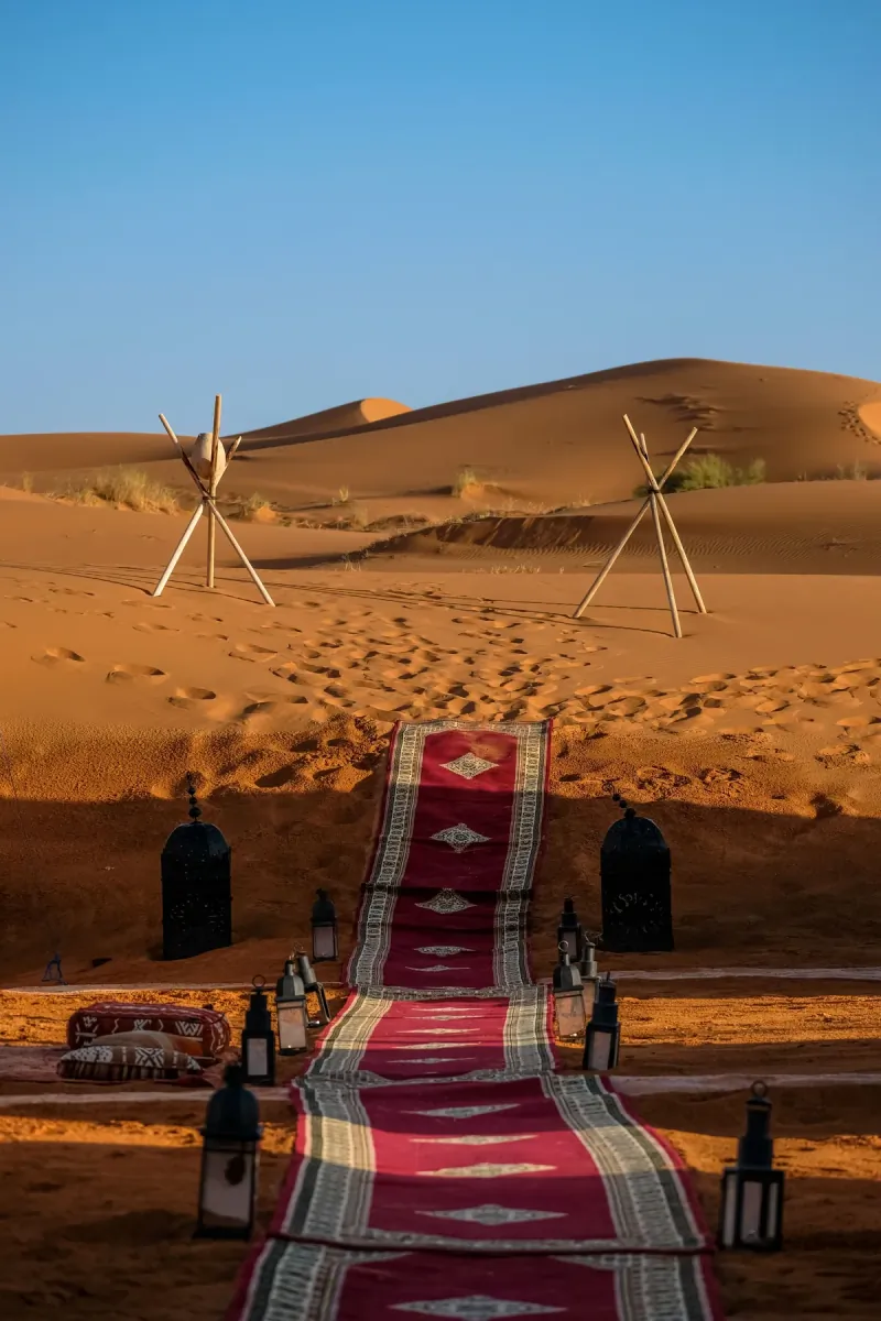Red Berber carpet laid across the sand leading to a desert camp at Merzouga, with lanterns on tall posts