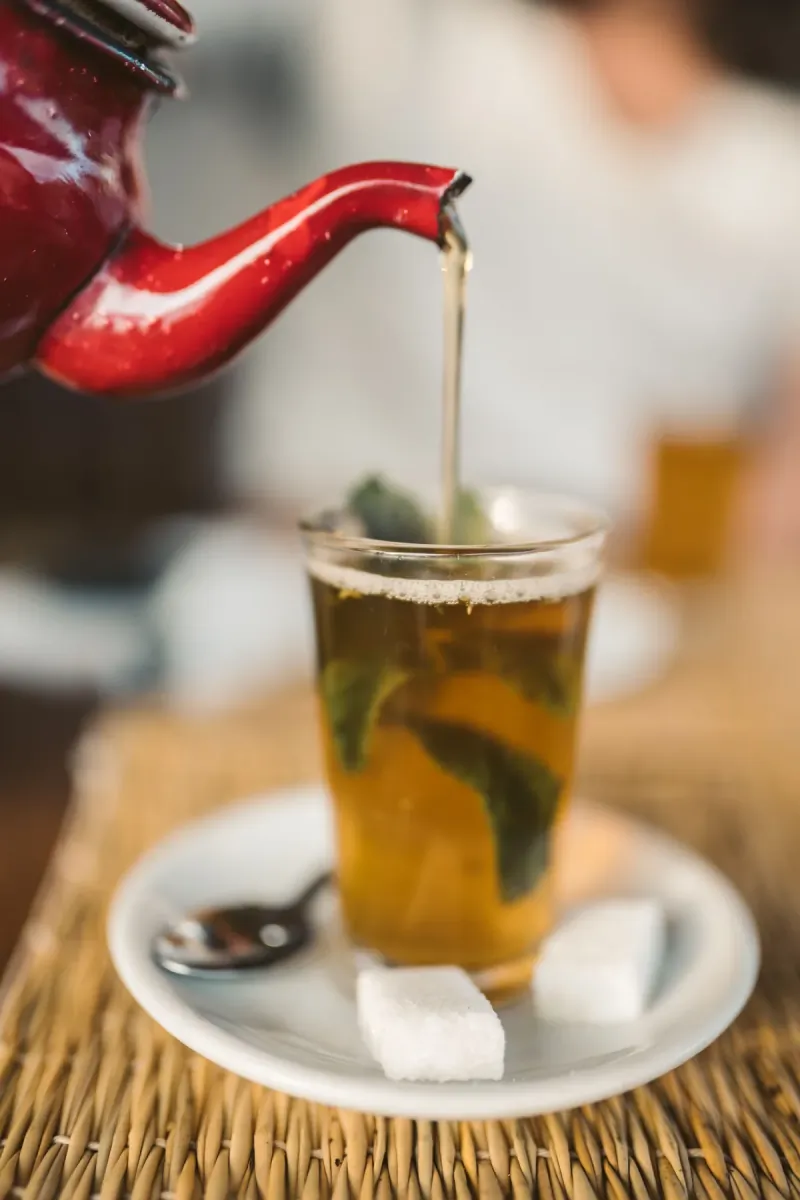 Moroccan mint tea being poured from a red teapot into a glass with fresh mint and sugar cubes