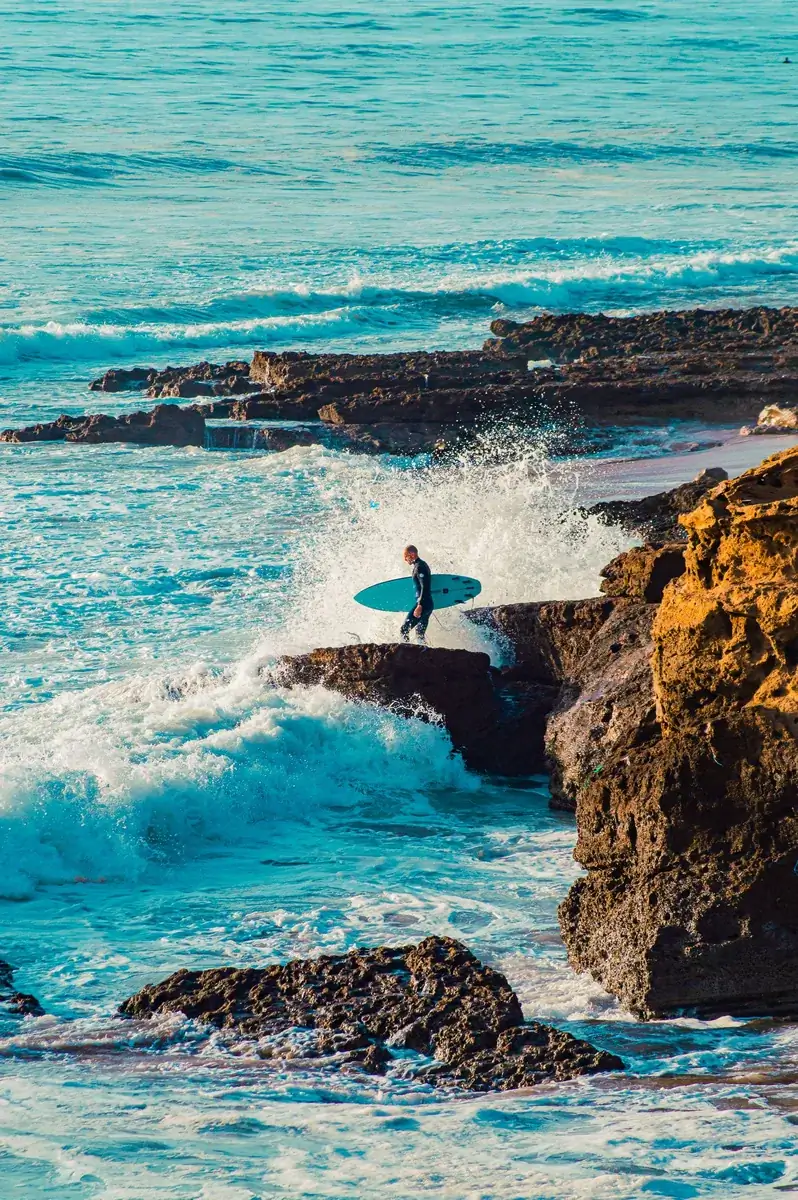 Surfer carrying a board across the rocks toward breaking Atlantic waves at Taghazout